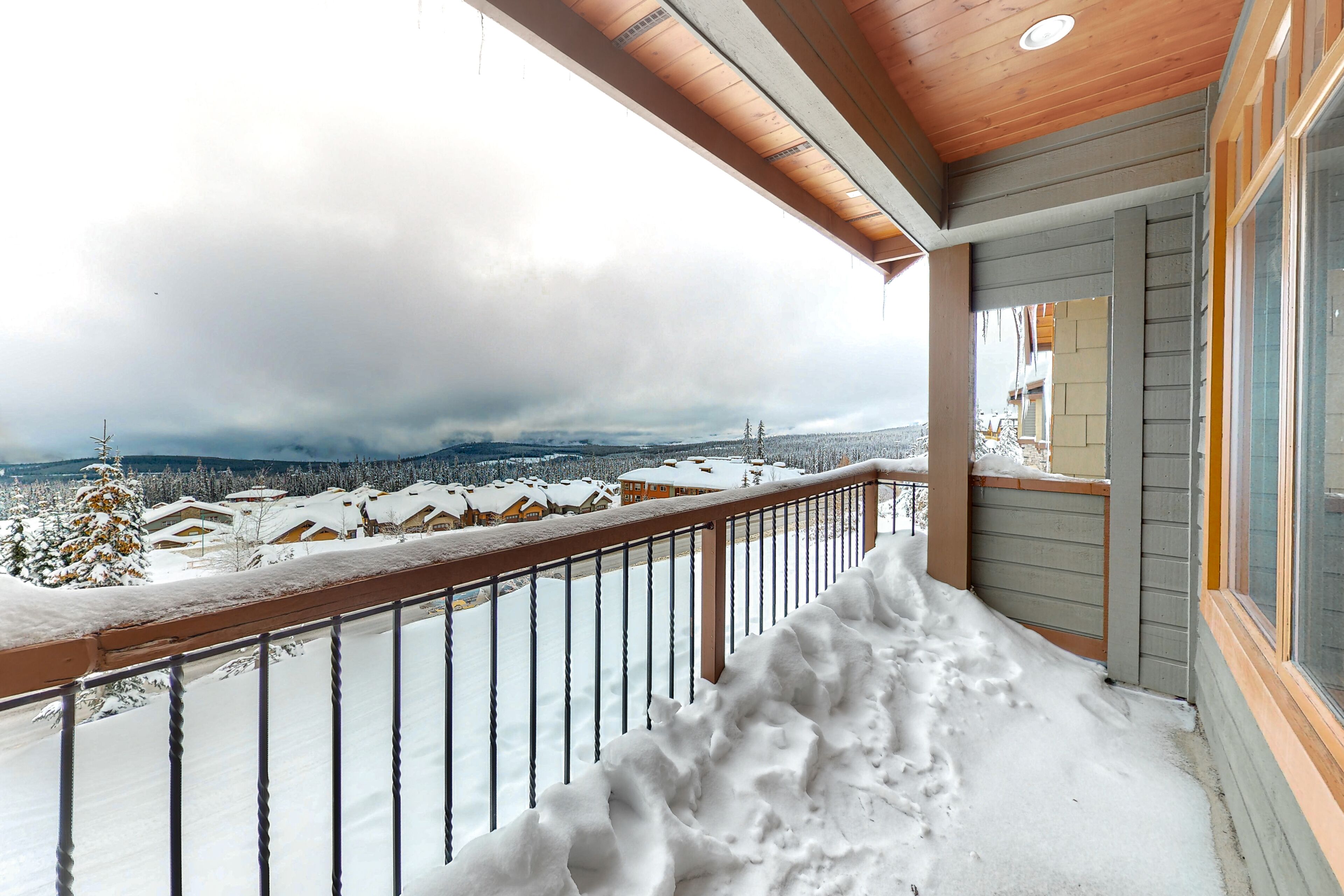 A snowy balcony with a wooden railing overlooking a snow-covered landscape and buildings.