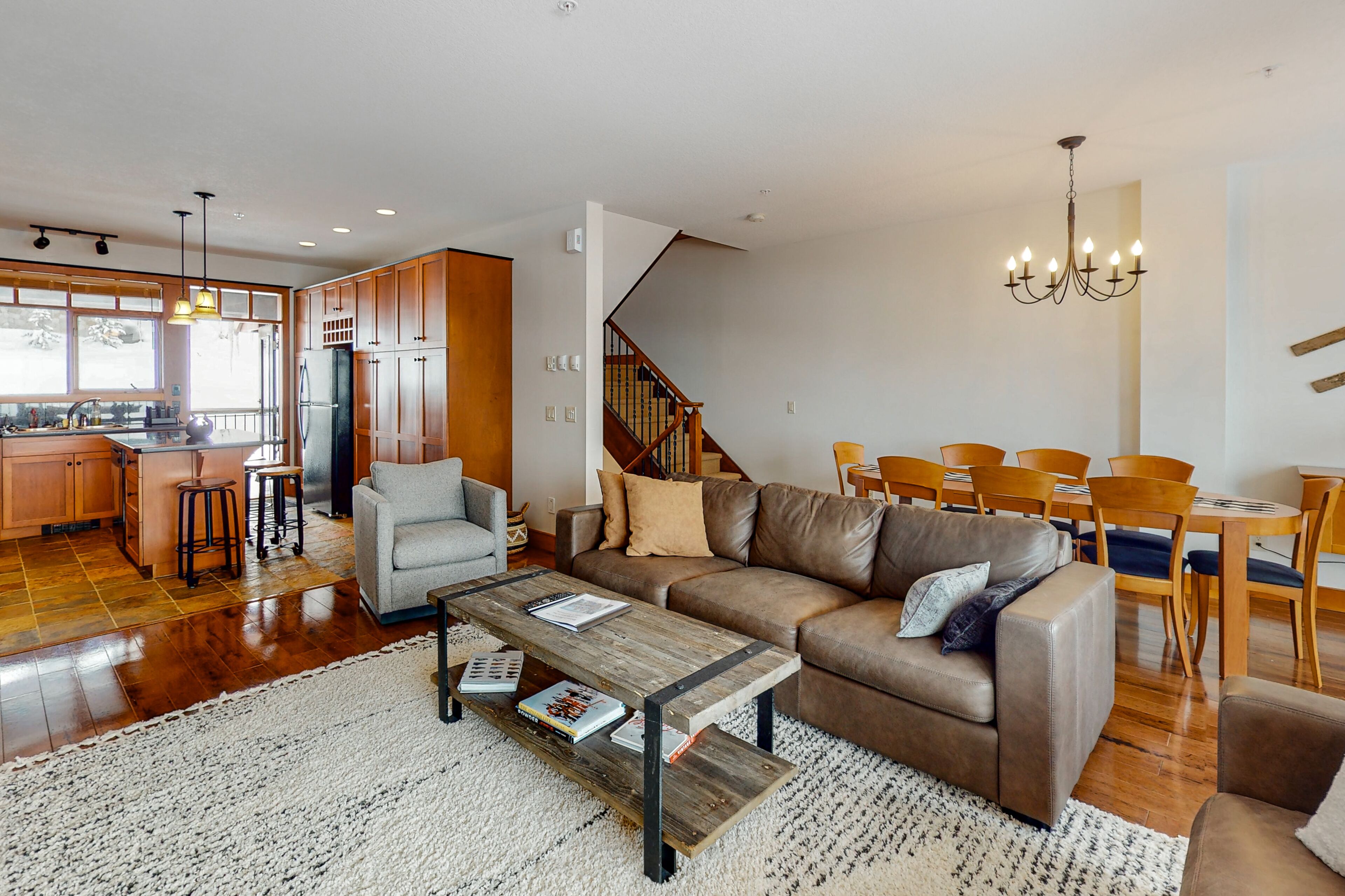A spacious living area with a large brown leather sofa, a wooden coffee table, and a dining table with chairs. A kitchen with wooden cabinets and a refrigerator is visible in the background.