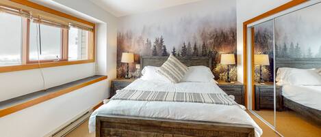A bedroom with a large bed, a window with blinds, a mirror, and a forest mural on the wall.