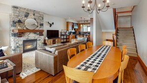 A spacious living area with a stone fireplace, a large dining table, and a kitchen in the background. - Blacksmith Lodge by Okanagan Premier (Beaverdell)