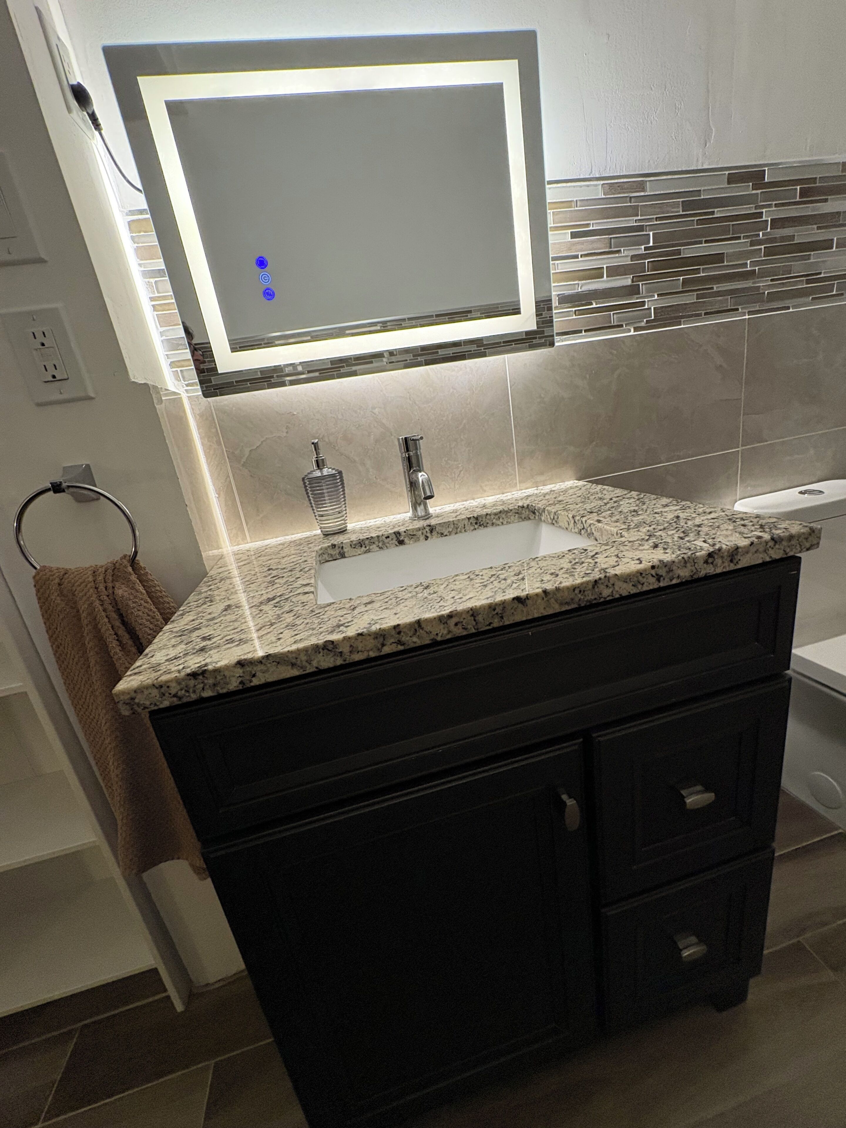 A bathroom with a granite countertop sink, a mirror with integrated lighting, a towel rack, and a tiled backsplash.