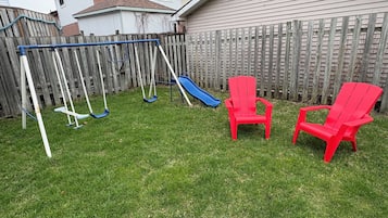 Un jardin avec une balançoire, deux chaises rouges et un toboggan bleu.