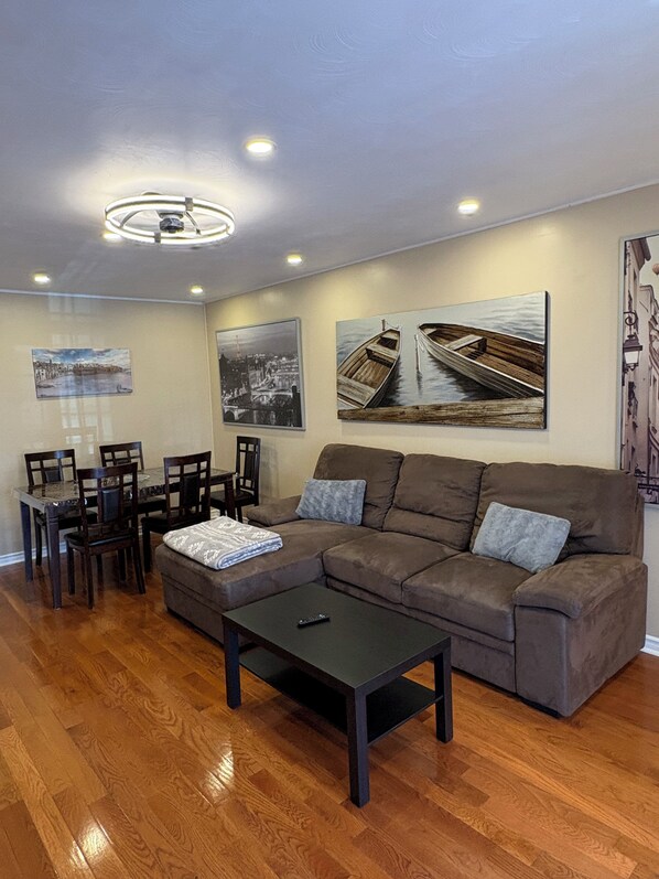 A living room with a brown sofa, a coffee table, a dining table with chairs, and artwork on the walls.