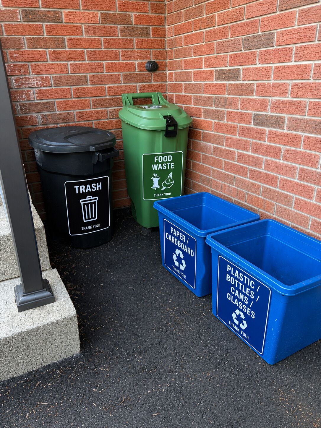 A trash bin, a green food waste bin, and two blue recycling bins against a brick wall.