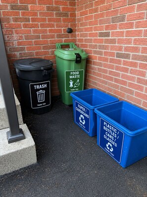 A trash bin, a green food waste bin, and two blue recycling bins against a brick wall.