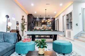 A modern living room with a pool table, a sofa, ottomans, and a dining area. - City View + Outdoor Projector With FirePit+PoolTbl+Golf+Dart+Golf+Grill+Sleeps16 (Dallas)