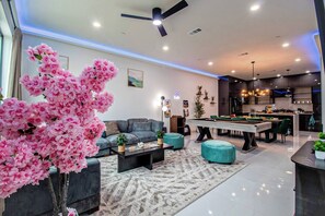 A modern living room with a sofa, coffee table, and ottomans, a pool table, and a kitchen area. - City View + Outdoor Projector With FirePit+PoolTbl+Golf+Dart+Golf+Grill+Sleeps16 (Dallas)