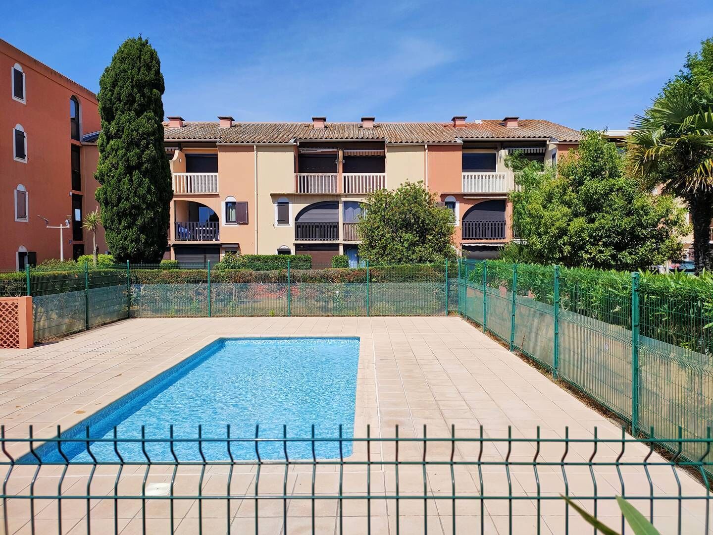 Una piscina con borde de baldosas, rodeada por una valla verde, y edificios de varios pisos con balcones al fondo.