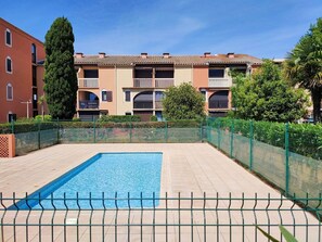 Ein Schwimmbecken mit gefliestem Deck, umgeben von einem grünen Zaun, im Hintergrund mehrstöckige Gebäude mit Balkonen.