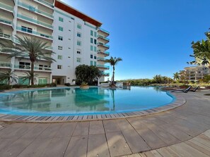 A swimming pool with a wooden deck, surrounded by palm trees and a multi-story building with balconies. - Depa con alberca infinity frente a la marina (Mazatlán)
