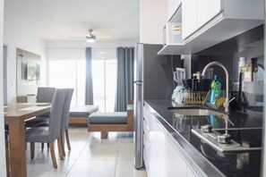 A modern kitchen with a black countertop, a built-in microwave, and a blender. There is a dining area with a table and chairs, and a large window with curtains. - Depa con alberca infinity frente a la marina (Mazatlán)