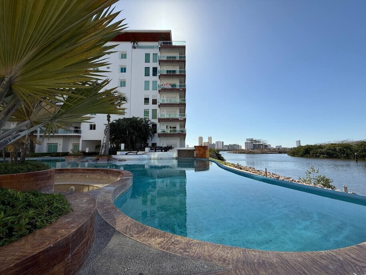 A swimming pool with a curved edge, surrounded by a landscaped area with plants and a palm tree. In the background, there is a multi-story building with balconies.