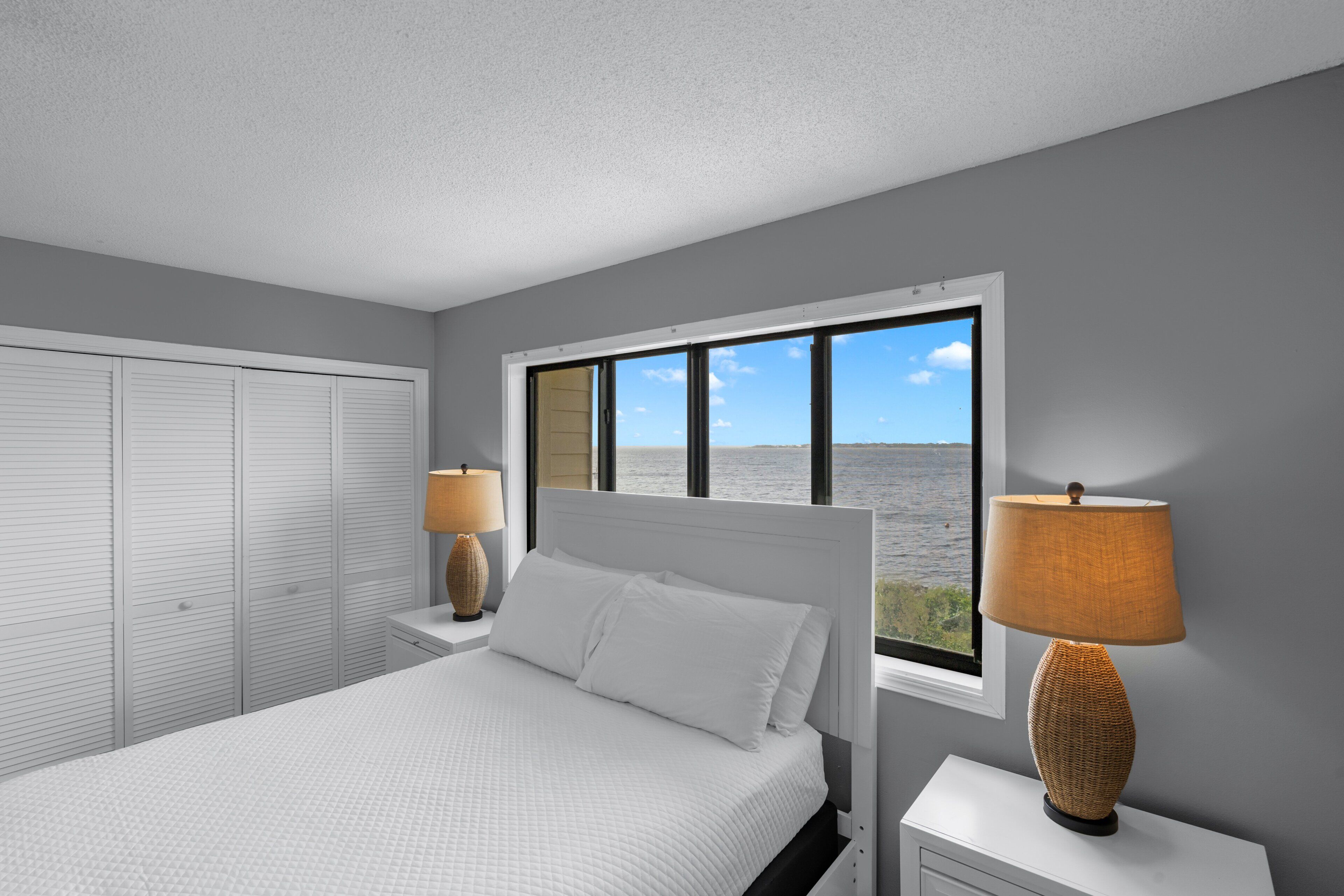 A bedroom with a bed, two nightstands, a window with a view of the sea, and a door with louvered shutters.
