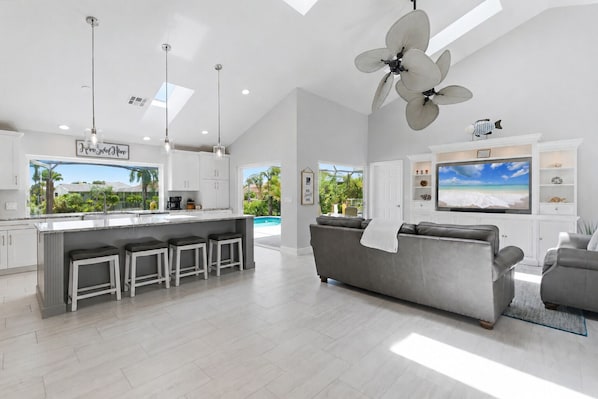 A modern kitchen with a bar area, a grey sofa, and a TV in a living area with a view of the outdoors. - Punta Gorda Private Pool Vacation Home (Punta Gorda)