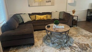 A living room with a brown sectional sofa, a round coffee table with a small plant, and a large abstract painting on the wall. - Day Dreams Begin at Bryant Denny Stadium 6 ppl (Tuscaloosa)