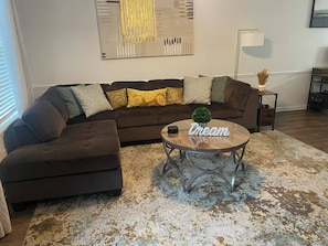 A living room with a brown sectional sofa, a round coffee table with a small plant, and a large abstract painting on the wall. - Day Dreams Begin at Bryant Denny Stadium 6 ppl (Tuscaloosa)