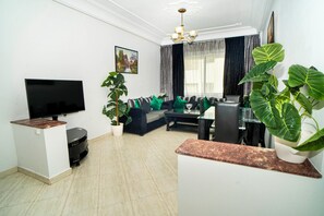 Ein Wohnzimmer mit Flachbildfernseher, einer Couch mit grünen Kissen, einem Couchtisch, einer Topfpflanze und einer Pendelleuchte.
