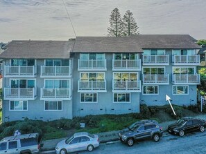 Un immeuble résidentiel à plusieurs étages, doté de balcons, avec des voitures garées et un arbre en arrière-plan.