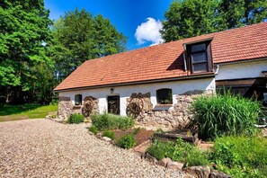 A rustic stone building with a red-tiled roof, a gravel driveway, and a garden with plants and a wooden fence.