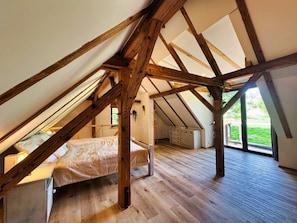 A loft bedroom with exposed wooden beams, a bed, a dresser, and a large window.