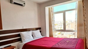 A hotel room with a bed, a window with curtains, an air conditioning unit, and a nightstand with a lamp. - PUKANTU  (Puno)