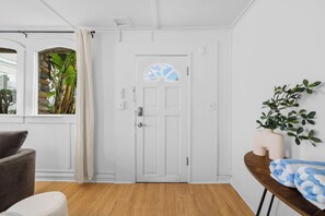 Ein Zimmer mit weißer Tür, einem Fenster mit Blick auf Grünflächen, einem braunen Stuhl, einem kleinen Tisch mit Pflanze und Handtuch.
