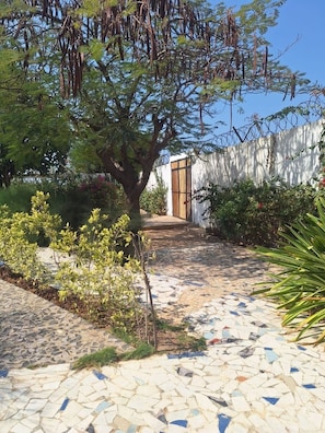 A garden pathway leading to a white building with a wooden door, surrounded by various plants and trees. - Blue Lagoon | Garden Floor (Warang)
