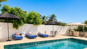 Ein Swimmingpool mit blauen Liegestühlen und Sonnenschirmen, umgeben von einer weißen Mauer und viel Grün.