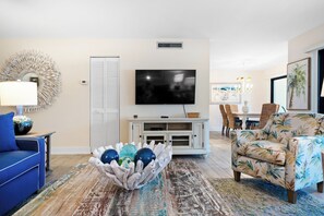 Ein Wohnzimmer mit Fernseher, einem blauen Sofa, einem gemusterten Sessel, einer Dekoschale und einem Essbereich mit Stühlen und Tisch.