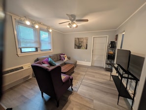 Un salon comprenant un canapé d’angle, un fauteuil foncé, une table basse, un ventilateur de plafond, un meuble pour la télévision et une porte.