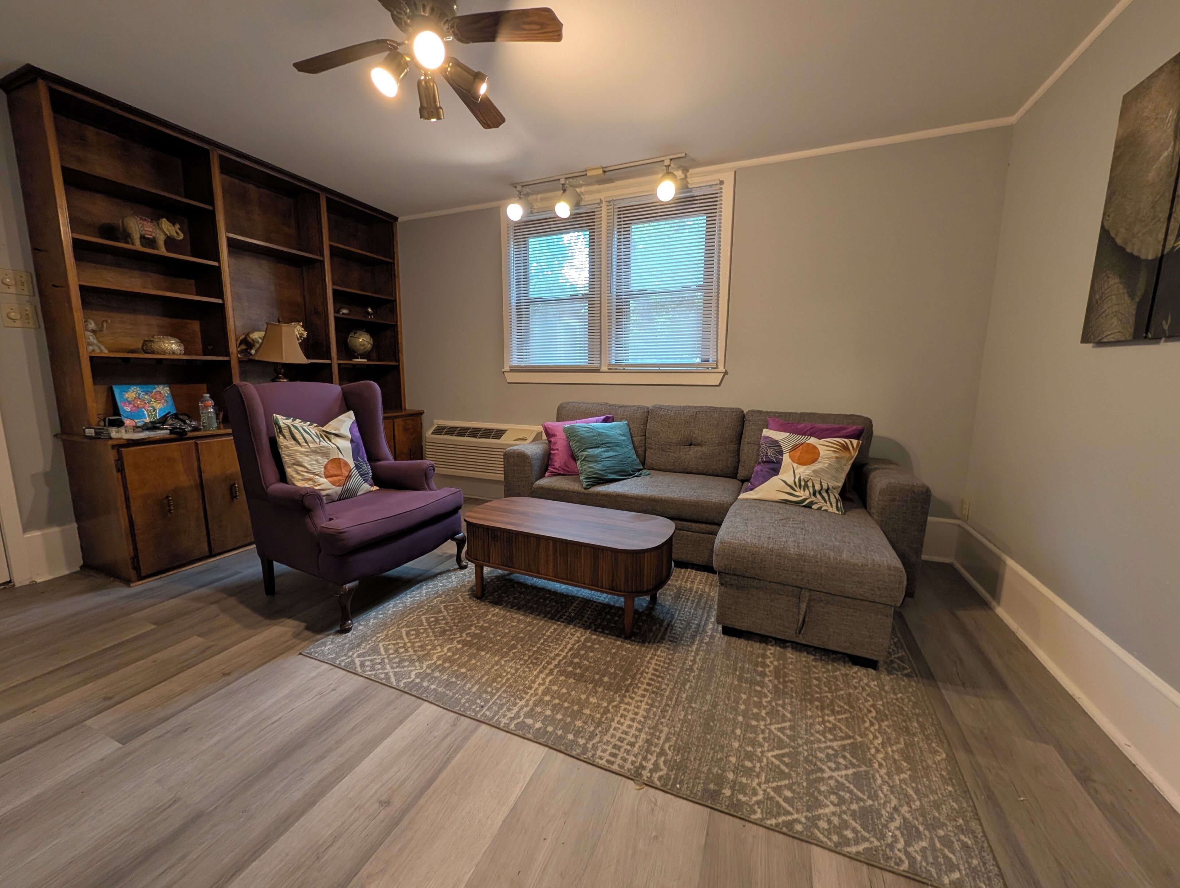 A living room with a purple armchair, a grey sofa, a wooden coffee table, and a patterned rug. There is a ceiling fan, a window with blinds, and a bookshelf.