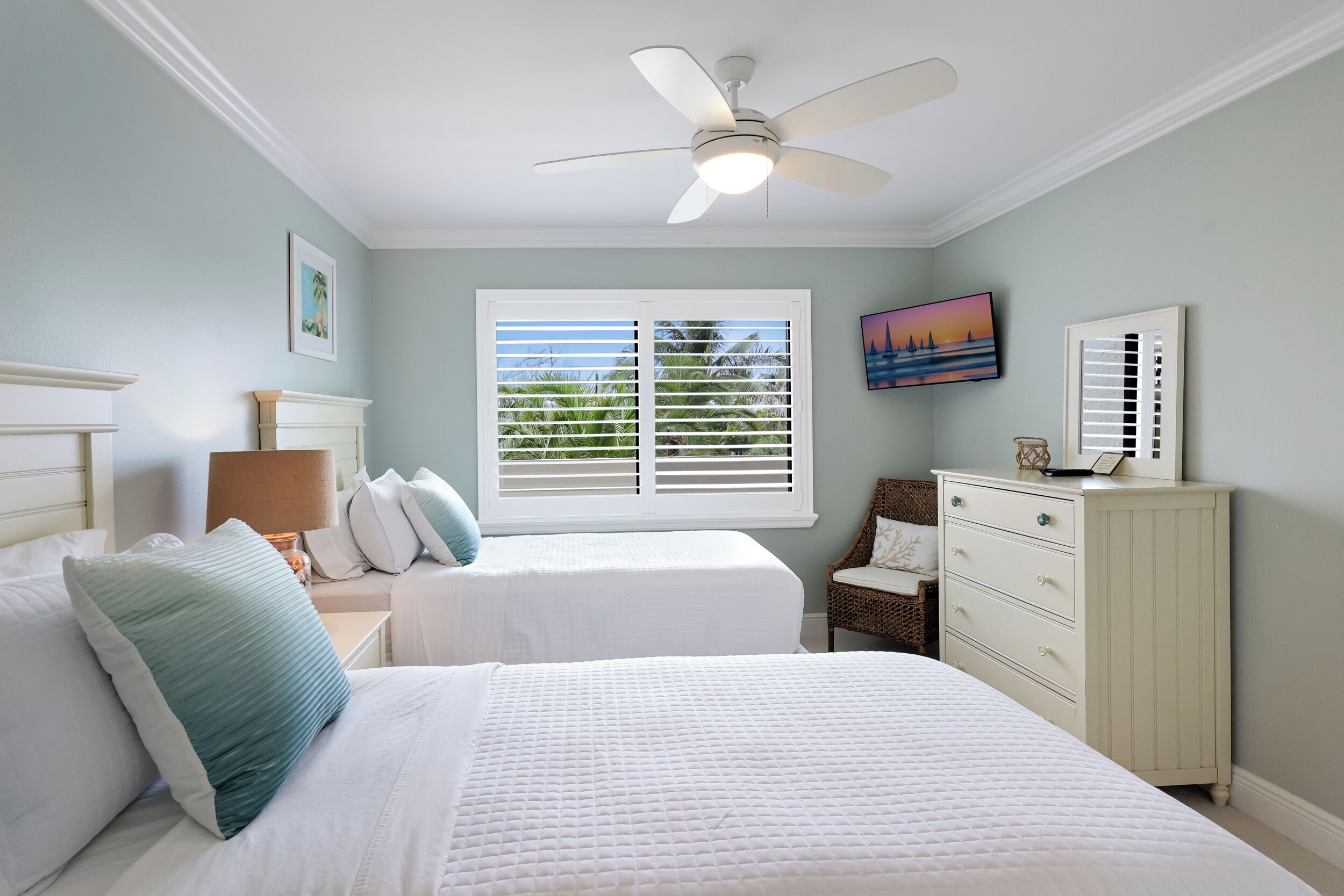 Quarto com duas camas, ventilador de teto, janela com persianas, televisão e cômoda.