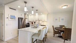 Une cuisine moderne avec des meubles blancs, un réfrigérateur en acier inoxydable, un éclairage suspendu et un coin repas avec des chaises en osier et une table en bois.