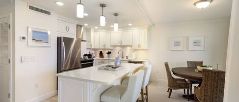 A modern kitchen with white cabinetry, a stainless steel refrigerator, pendant lighting, and a dining area with wicker chairs and a wooden table.