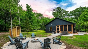 Eine moderne Holzhütte mit Feuerstelle und Adirondack-Stühlen in bewaldeter Landschaft.