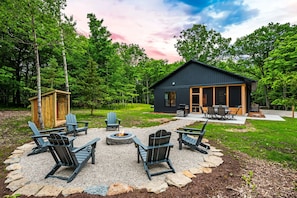 Une cabane moderne avec un foyer et des fauteuils Adirondack, située dans un cadre forestier.