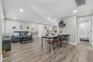 Ein modernes Wohnzimmer mit Sofa, Couchtisch, Esstisch und Stühlen. Es gibt eine Küchenzeile mit Kühlschrank und Mikrowelle.