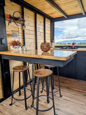 Una cocina con encimera de madera, taburetes, una rueda decorativa y vistas a montañas.