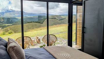 A bedroom with a large window, a bed, a chair, and a view of a mountainous landscape.
