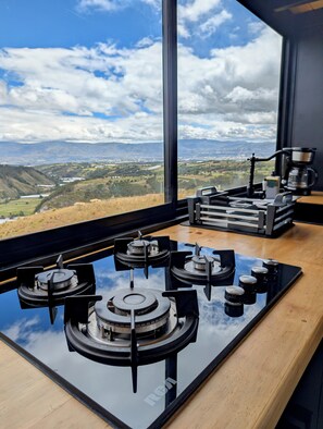 Una cocina con estufa a gas, cafetera y vistas a un paisaje montañoso a través de un ventanal grande.
