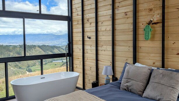 Un dormitorio con un ventanal grande, una bañera, una cama con almohadas y un techo de madera.