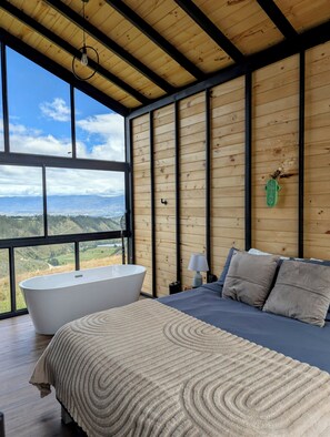 Un dormitorio con un ventanal grande, una bañera, una cama con almohadas y un techo de madera.