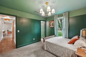 Ein Schlafzimmer mit Bett, Nachttisch, einem Fenster mit Vorhängen, einer Tür und einem Flur, der in ein weiteres Zimmer führt.