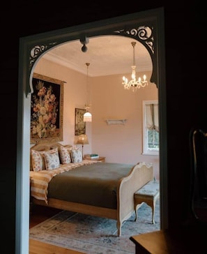A bedroom with a bed, a chandelier, a painting, and a window.
