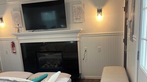 A room with a fireplace, a television mounted above it, a bench, and a bed with a patterned bedspread. - Highlands cottage across the street from Proving Ground and Twin Lights Marina (Highlands)