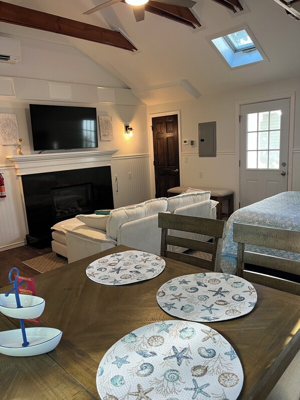 A living room with a fireplace, a television, a dining table with placemats, a ceiling fan, and a skylight.