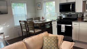 A modern kitchen with a built-in microwave, stove, and cabinets. There is a dining area with a table and chairs, and a living area with a sofa and cushions. - Highlands cottage across the street from Proving Ground and Twin Lights Marina (Highlands)