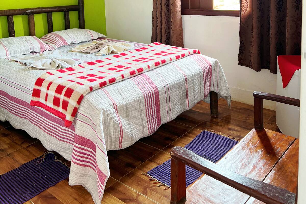 A room with a bed, wooden headboard, a window with curtains, and a wooden chair.