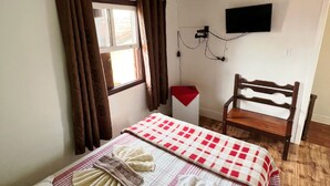 A neatly made bed with a checkered blanket, a wooden chair, a television mounted on the wall, and a window with curtains. - Pousada Cantinho Gostoso (Ouro Preto)