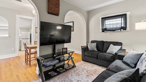 A living room with a flat-screen TV, grey sofa, and a dining area with a wooden table and chairs.